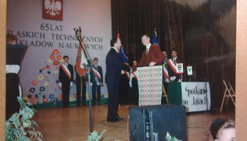 Dyrektor Szkoły wita na scenie gościa, z tyłu poczty sztandarowe