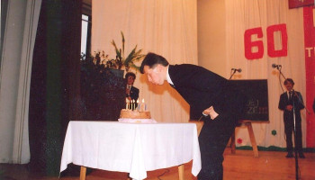 scena w auli, na stole nakrytym białym obrusem urodzinowy tort; Pan Dyrektor zdmuchuje świeczki na torcie