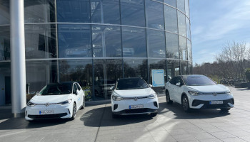 Przeszklony budynek, przed nim trzy białe samochody elektryczne Volkswagen