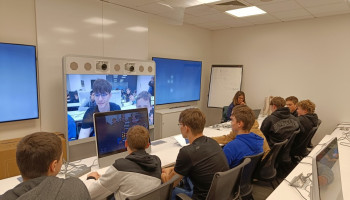 Uczniowie podczas wykładu, przed nimi bardzo duże ekrany, na których widoczne są twarze uczniów