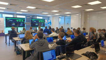 Uczniowie siedzący przy stołach z laptopami, przed nimi dwa ekrany LCD