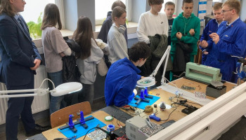 Pracownia montażu układów elektronicznych 3 uczniów w fartuchach ochronnych (niebieskie) prezentują grupie 7 odwiedzających leżące na stanowisku materiały dydaktyczne do lutowania
