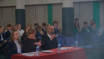 Jury konkursu w składzie pani wicedyrektor, nauczyciel wf i członkini Zarządu SU przy stole prezydialnym podczas występów, za jury widoczna publiczność-uczniowie szkoły