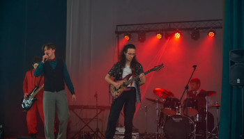 Występ - występujący zespół: z lewej wokalista, za nim gitarzysta, w środku gitarzysta z gitara elektryczną, po prawej grający na perkusji, oświetlenie w kolorze czerwonym