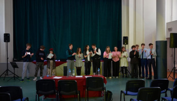 Wyróżnieni i uczestnicy (14 osób) stoją przed sceną w auli, finał konkursu, wyróżnieni z dyplomami, wszyscy biją brawo, kurtyna zasłonięta w tle, jury opuściło stół prezydialny