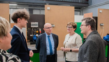 w pracowni elektrycznej, uczeń w garniturze referuje na temat zajęć pani minister i pani poseł; pani dyrektor z prawej, nauczyciel przedmiotów elektrycznych w środku