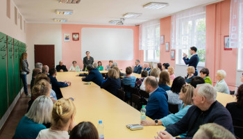 pokój nauczycielski, spotkanie, ujęcie perspektywiczne grona siedzącego przy stołach po dwóch stronach, w centrum pani minister w trakcie przemowy