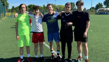 drużyna 1D – grupa 5 zawodników, stroje sportowe, pozycja stojąca, objęci za ramiona, w tle boisko 