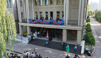 powitanie gości, wejście do budynku szkoły, stoją: zaproszeni, pani dyrektor; nauczyciel u dołu schodów prezentuje przybyłych, niżej stoi pani wicedyrektor, po lewej para uczniów w strojach śląskich, na balkonie szkoły udekorowanym niebieskimi i czerwonymi balonami zespół muzyczny (5 uczniów z instrumentami)