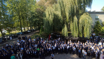 społeczność szkolna zebrana na uroczystości – bardzo liczna grupa uczniów, nauczyciele – stoją frontem do szkoły; w tle drzewa rosnące na skwerze przed szkołą