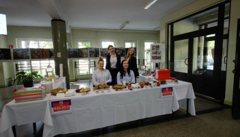 stoisko kiermaszu z nazwą „Kołocz” w holu szkoły; 4 uczennice: 2 siedzą – białe bluzki, 2 stoją – ubrane w granatowe mundurki – za stołem nakrytym białym obrusem z różnymi ciastami