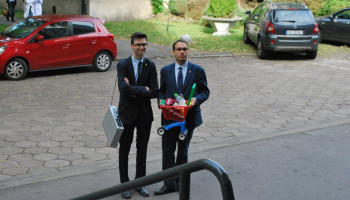 2 uczniów: jeden z urządzeniem elektronicznym przewieszonym przez ramię, drugi trzyma jako plecak wózeczek na zakupy 