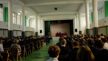 aula szkolna: uczniowie uczestniczący w debacie, widok na scenę – za stołem prezydialnym trzej siedzący kandydaci, jeden przy mównicy